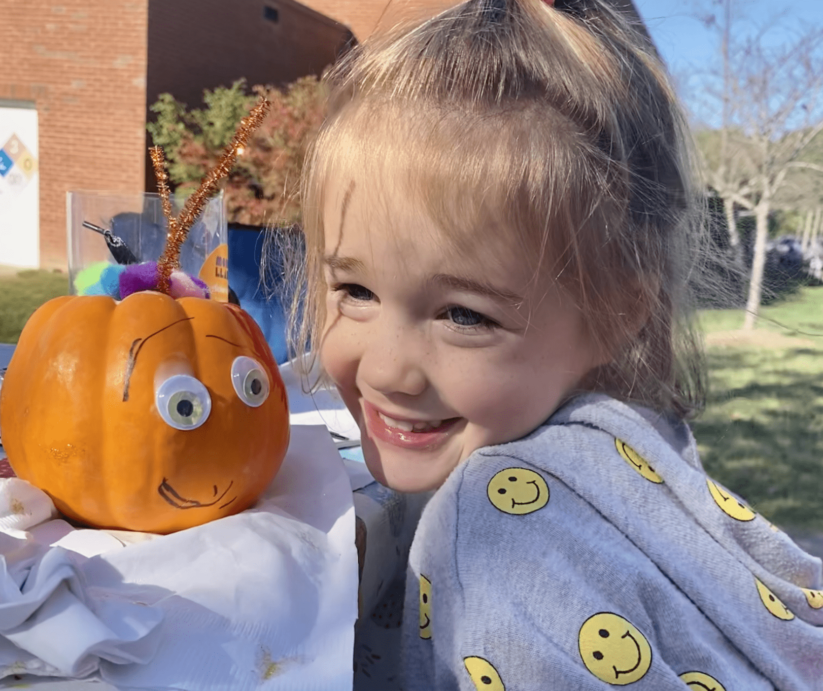 Picture of little girl smiling next to an orange pumpkin with googly eyes and a smile on it