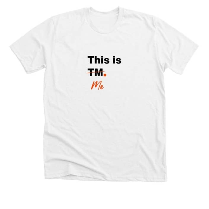 White t-shirt with This is TM in black text. TM is crossed out with orange ink and beneath is the word Me in orange text.