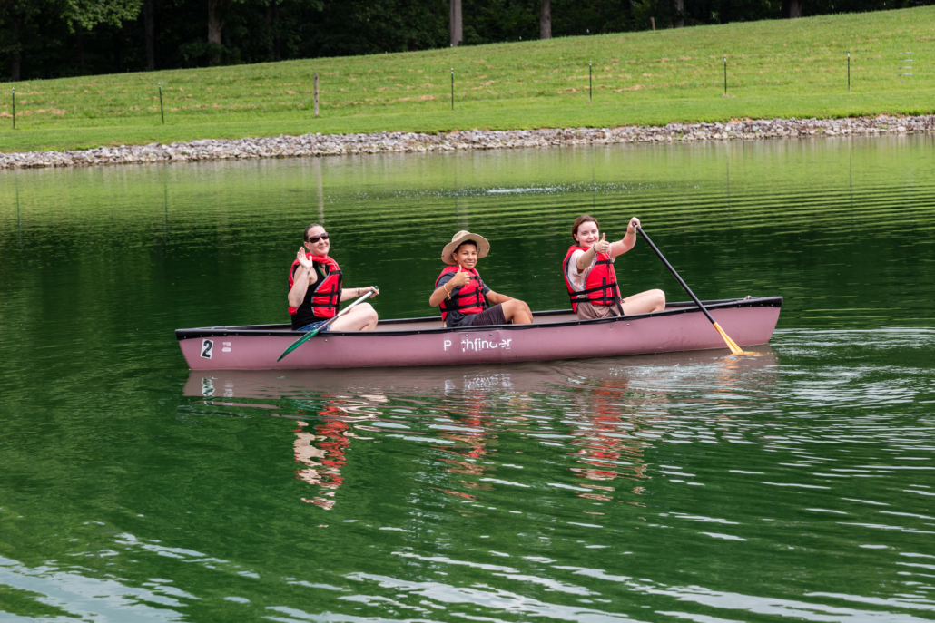 3 people with orange life vests sitting in a pink canoe in a greenish lake all smiling at the camera