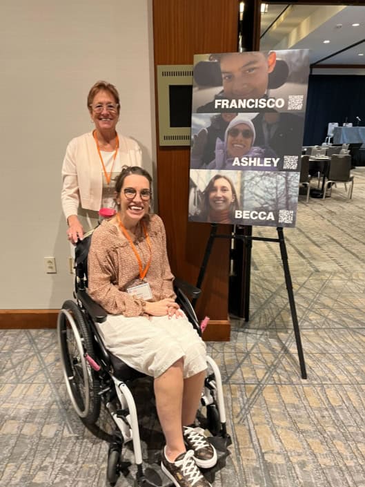 Photo of Ashley, seated in her chair, with her mother Mary standing behind her. They're next to a poster with three community members highlighted: Francisco, Ashley, and Becca.